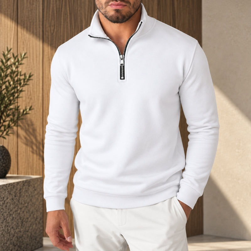 A man wearing a white long-sleeve quarter-zip pullover sweater with a black zipper pull, paired with white pants, set against a modern wooden and concrete background.
