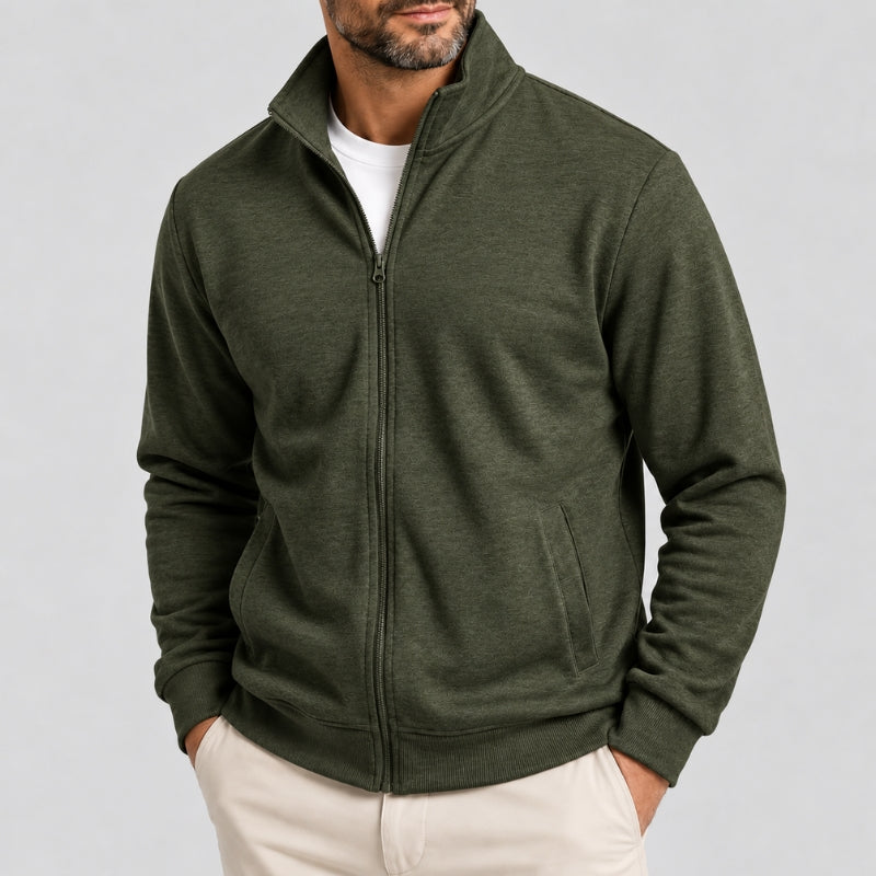 A man wearing an olive green full-zip fleece jacket over a white t-shirt and cream pants, shown from the chin down against a light gray studio background.