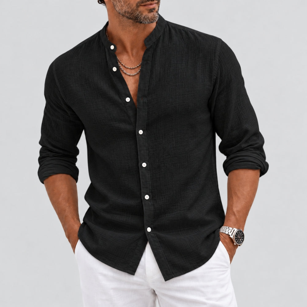 A man wearing a black long-sleeve linen shirt with white buttons and white pants, shown from the chin down against a light gray background.