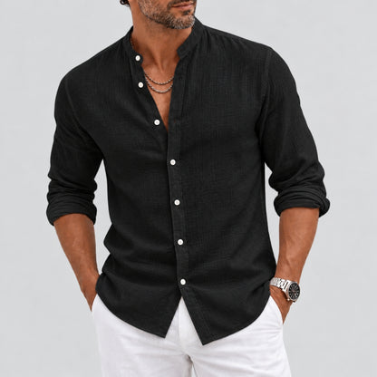 A man wearing a black long-sleeve linen shirt with white buttons and white pants, shown from the chin down against a light gray background.