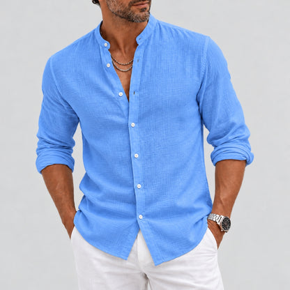 A man wearing a light blue, long-sleeve linen button-down shirt with rolled-up sleeves and white pants, shown from the chin down against a light gray background.