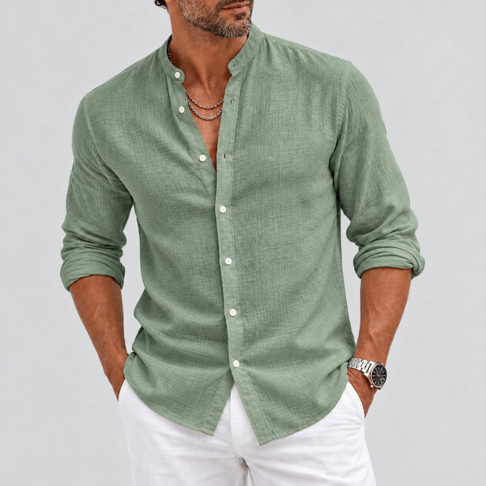A man wearing a sage green long-sleeve linen button-up shirt with white pants, standing against a light gray studio background.