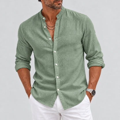 A man wearing a sage green long-sleeve linen button-up shirt with white pants, standing against a light gray studio background.