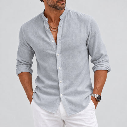 A man wearing a light gray linen long-sleeve button-down shirt with white pants, standing against a light gray studio background.
