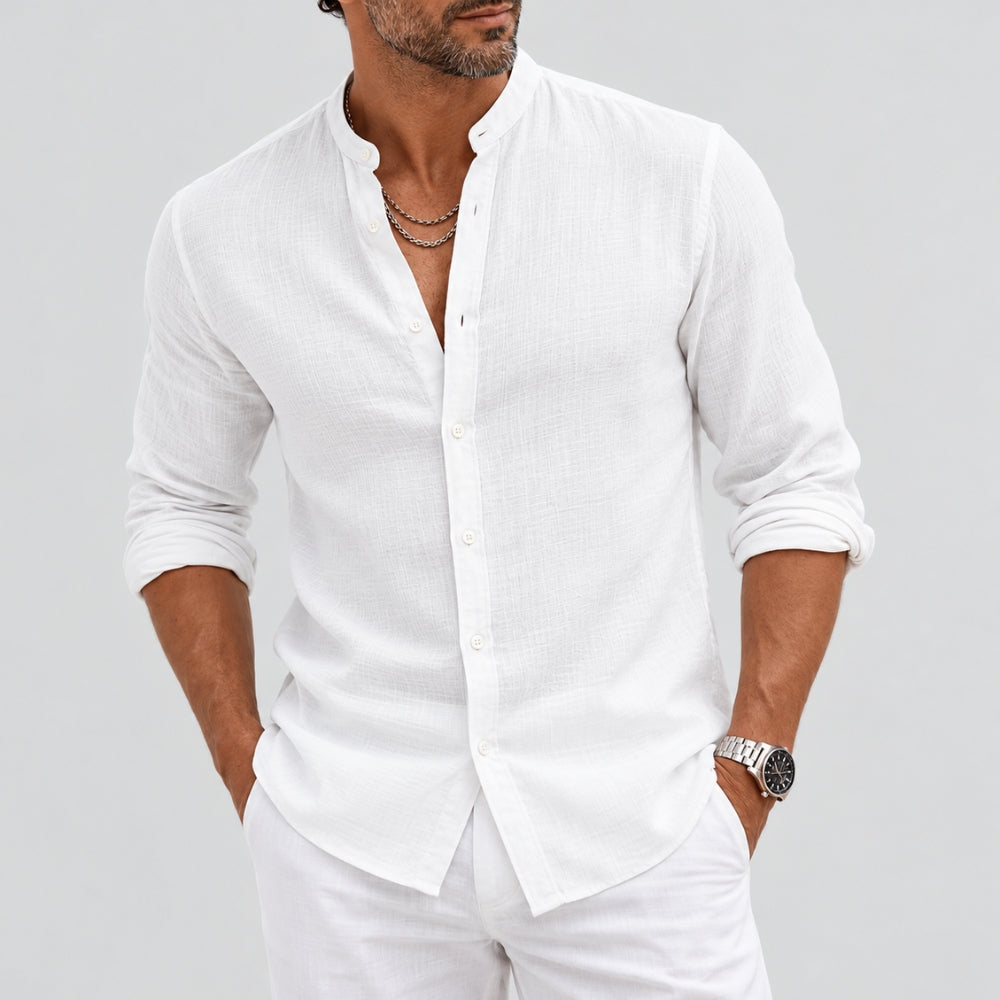 A man wearing a white long-sleeve linen button-down shirt with rolled-up sleeves and white trousers, standing against a light gray studio background.