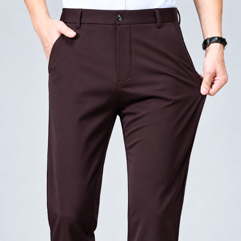A close-up of a person wearing dark brown trousers, holding the sides of the legs to demonstrate flexibility and comfort. The person is cropped from the waist down, with one hand in a pocket and the other pulling the fabric outward.