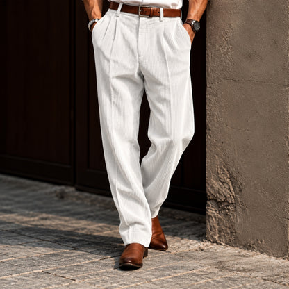 A person wearing white pinstriped trousers with a brown belt and brown leather shoes, standing outdoors against a dark wooden door and textured wall.