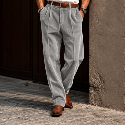 A person wearing light gray pinstriped corduroy trousers with a brown leather belt and brown leather shoes, standing outdoors against a dark wooden door and textured wall.