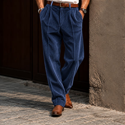 A person wearing blue corduroy trousers with a brown belt and brown leather shoes, standing outdoors against a dark wooden door and textured wall.