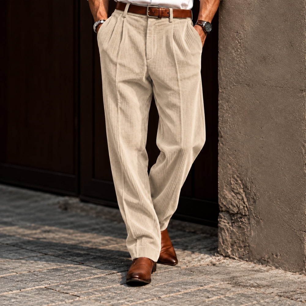 A person wearing beige corduroy trousers with a brown belt and brown shoes, standing outdoors against a dark wooden door and textured wall.