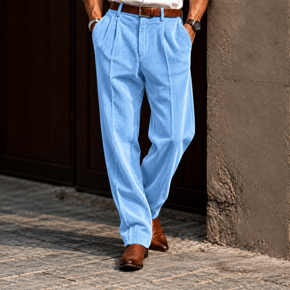 A person wearing light blue pinstriped trousers with a brown belt and brown leather shoes, standing outdoors against a dark wooden door and textured wall.