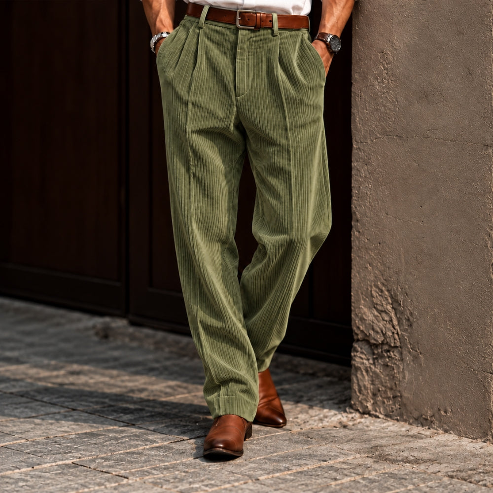 A person wearing olive green corduroy trousers with a brown belt and brown leather shoes, standing on a cobblestone street against a dark wooden door and textured wall.