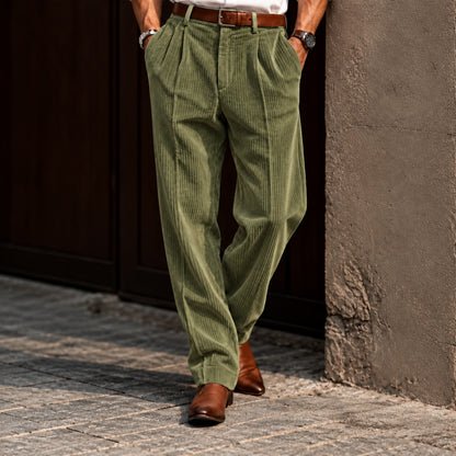 A person wearing olive green corduroy trousers with a brown belt and brown leather shoes, standing on a cobblestone street against a dark wooden door and textured wall.