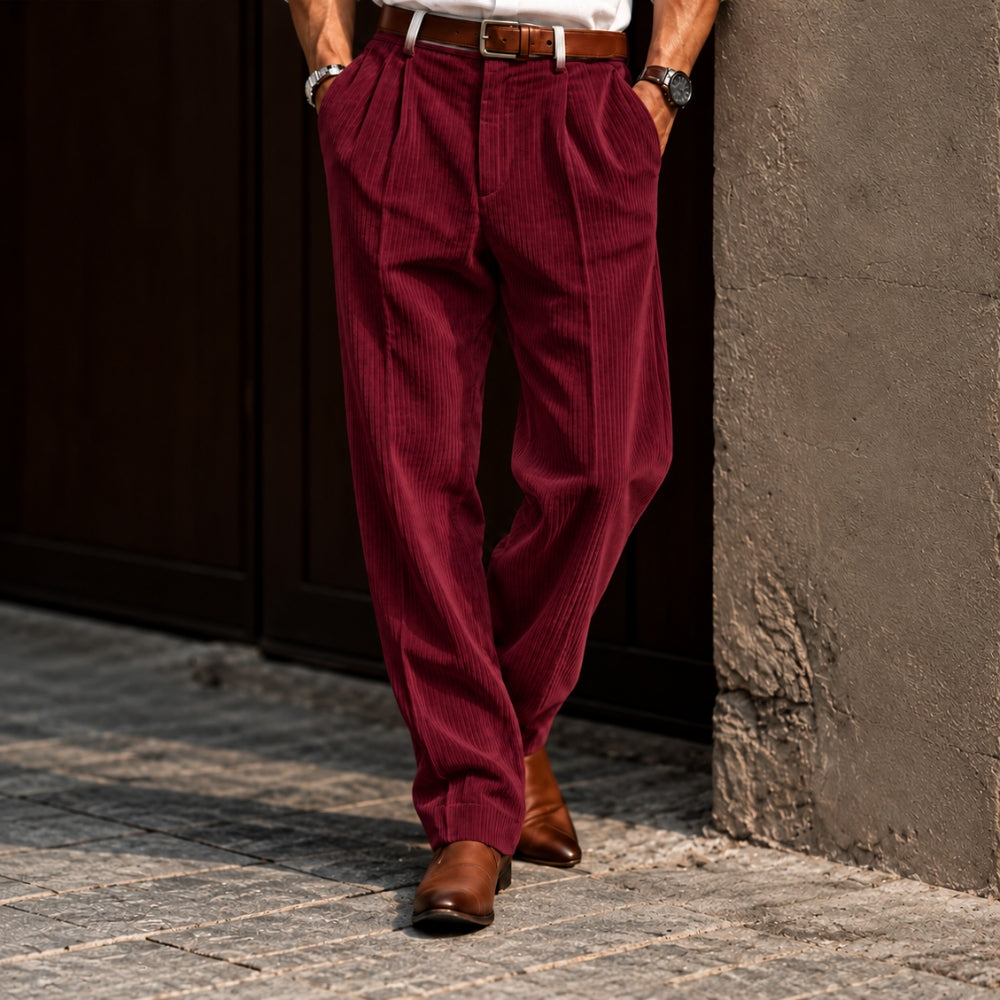 A person wearing pleated corduroy trousers in a deep burgundy red, styled with a white shirt, brown belt, and brown leather shoes, standing on a cobblestone street next to a textured wall.