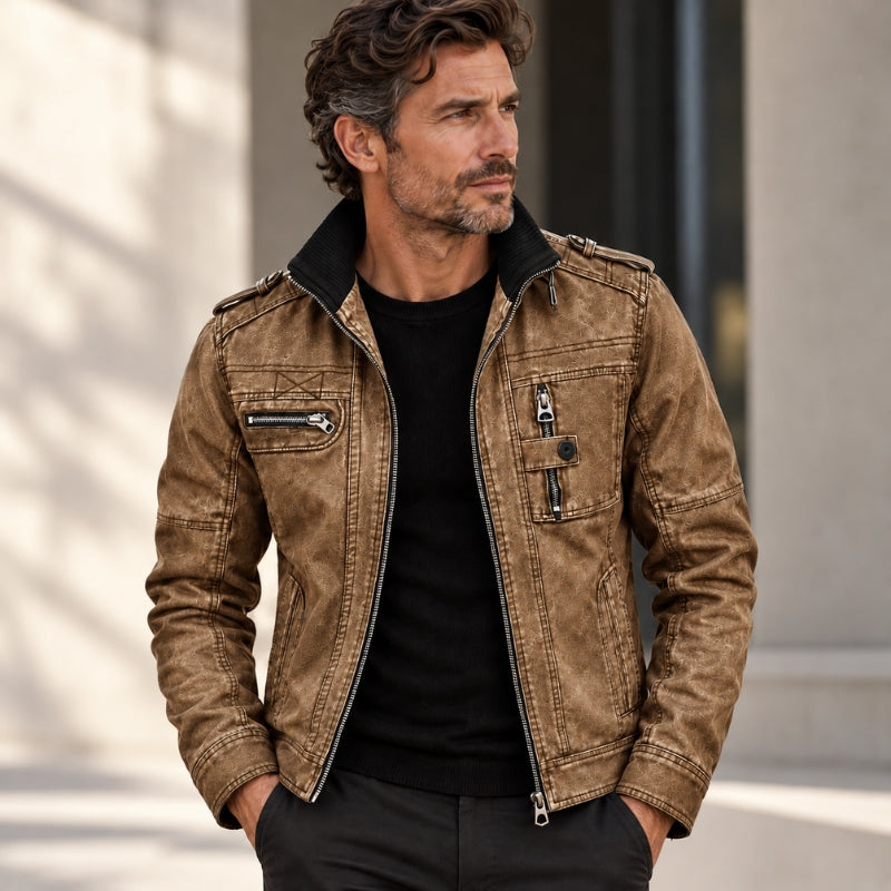 A man wearing a brown distressed faux-leather motorcycle jacket with a black collar, styled over a black t-shirt and dark pants, standing outdoors against a blurred beige building background.