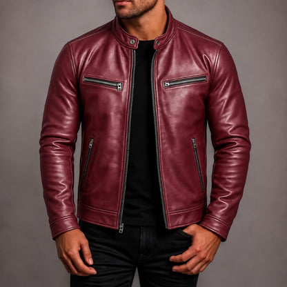 A man wearing a burgundy leather motorcycle jacket with silver zippers over a black shirt and black pants, photographed against a neutral gray studio background.