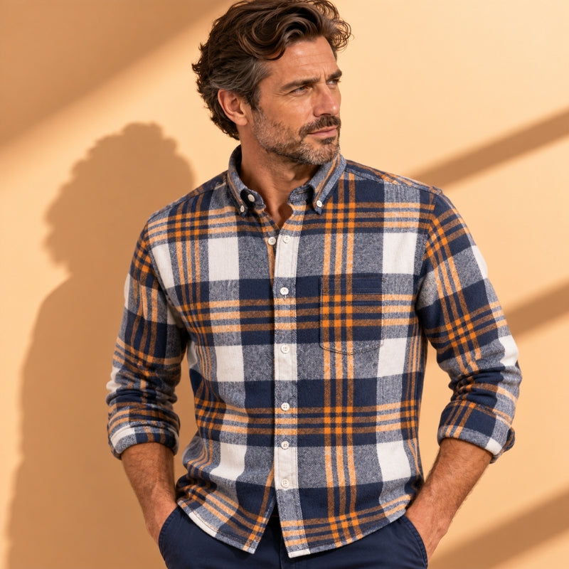A man wearing a long-sleeve button-down plaid shirt in navy blue, white, and orange, paired with navy pants, standing against a warm beige background with soft diagonal shadows.