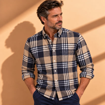 A man wearing a long-sleeve plaid button-down shirt in navy, beige, and white, paired with navy trousers, standing against a warm beige wall with soft shadows.
