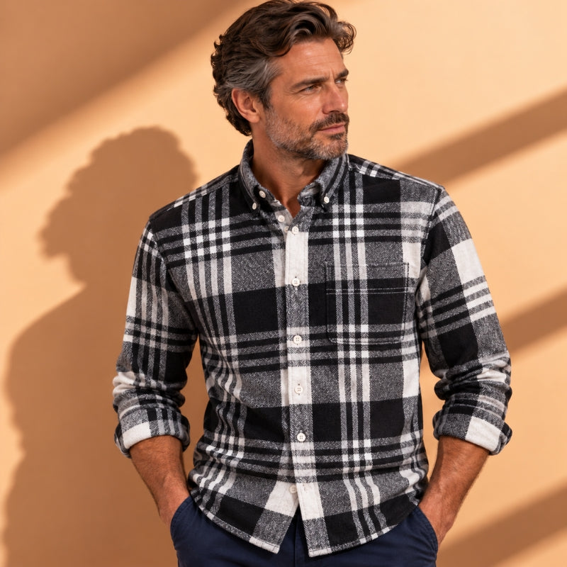A man wearing a black, white, and gray plaid button-down shirt, standing against a warm beige background with soft shadows.