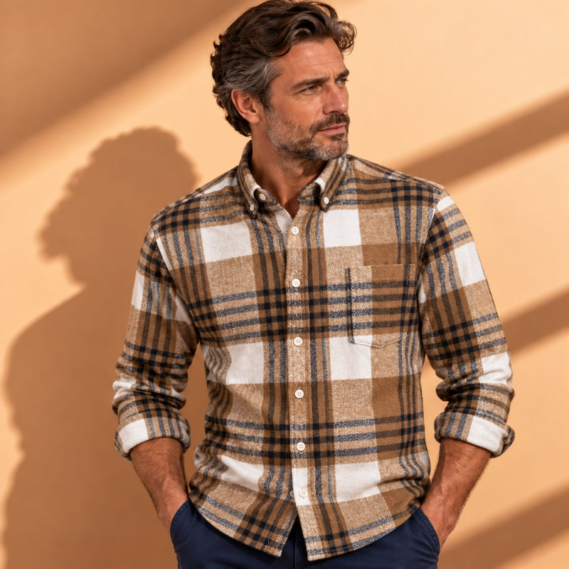 A man wearing a brown, beige, and white plaid button-down shirt, standing against a warm beige wall with soft shadows.