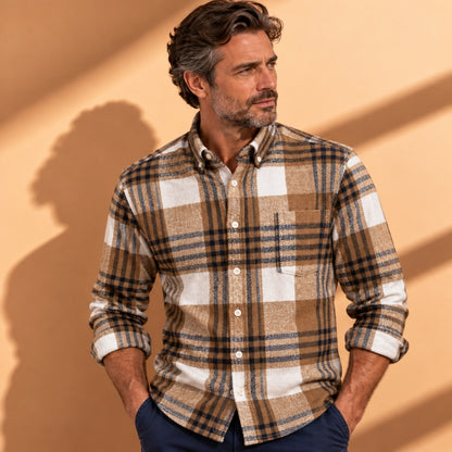 A man wearing a brown, beige, and white plaid button-down shirt, standing against a warm beige wall with soft shadows.