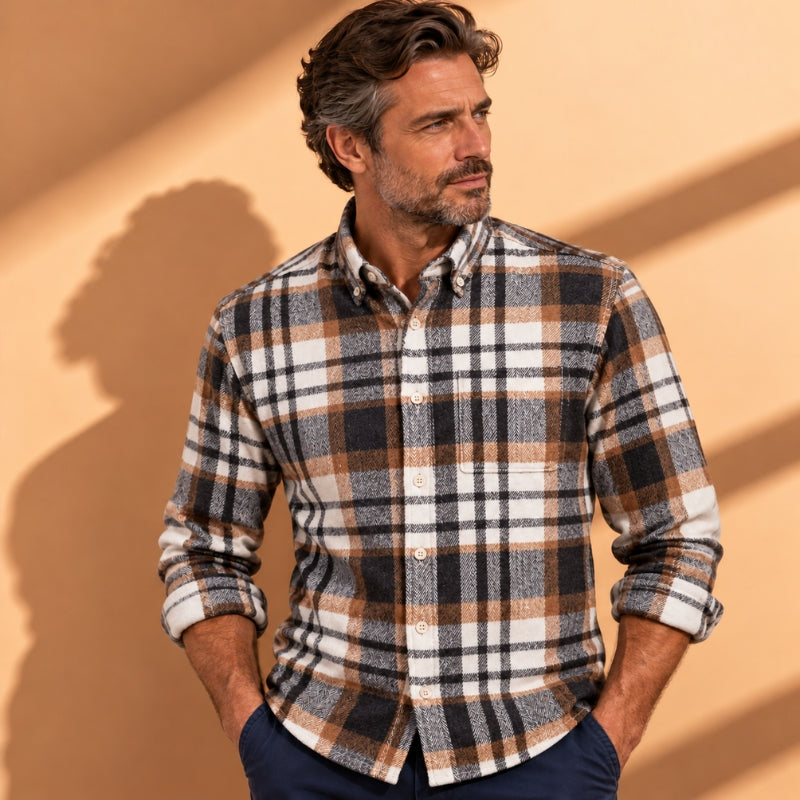 A man wearing a long-sleeve button-down plaid shirt in brown, black, and white, paired with navy blue pants, standing against a warm beige wall with soft shadows.