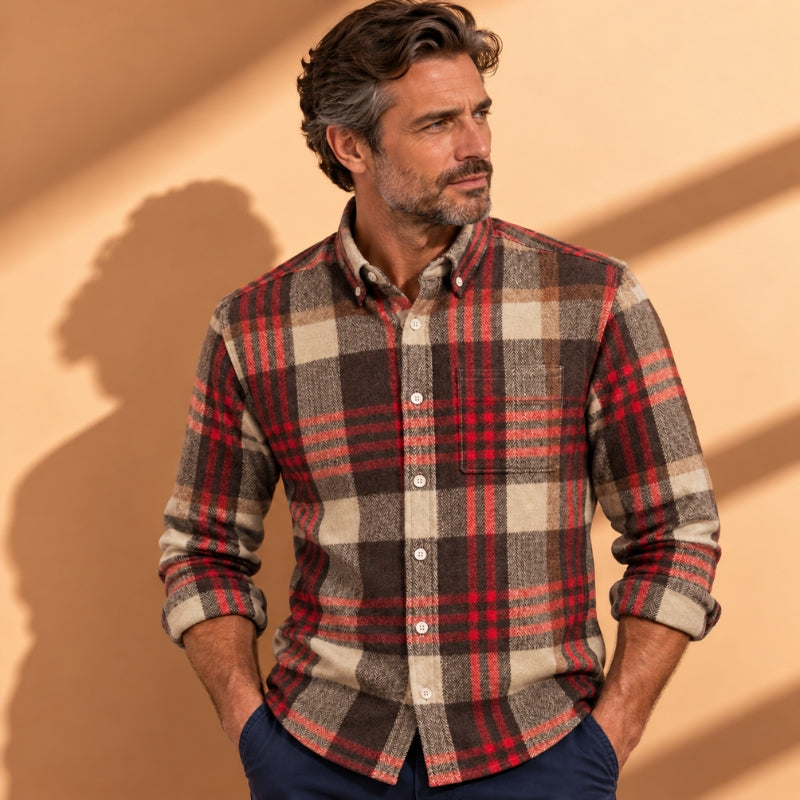 A man wearing a long-sleeve button-down plaid flannel shirt in red, brown, and beige, paired with navy blue pants, standing against a warm beige wall with soft shadows.