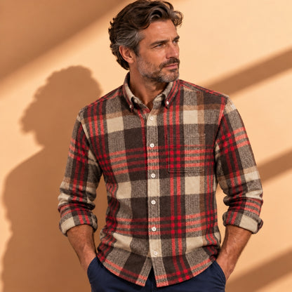 A man wearing a long-sleeve button-down plaid flannel shirt in red, brown, and beige, paired with navy blue pants, standing against a warm beige wall with soft shadows.