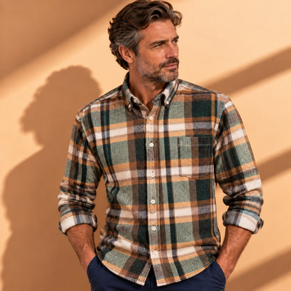 A man wearing a long-sleeve button-down plaid flannel shirt in green, brown, and beige tones, paired with navy pants, standing against a warm beige wall with soft shadows.