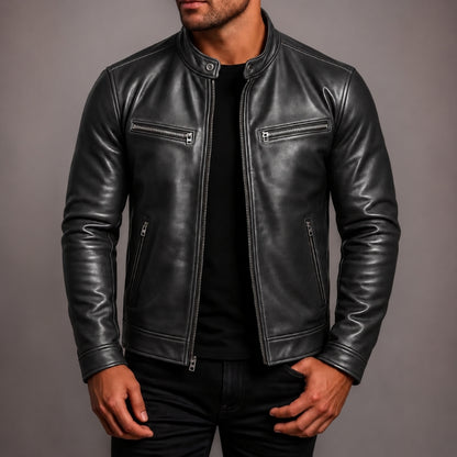 A man wearing a black leather jacket with silver zippers over a black shirt, shown from the chin down against a plain gray studio background.