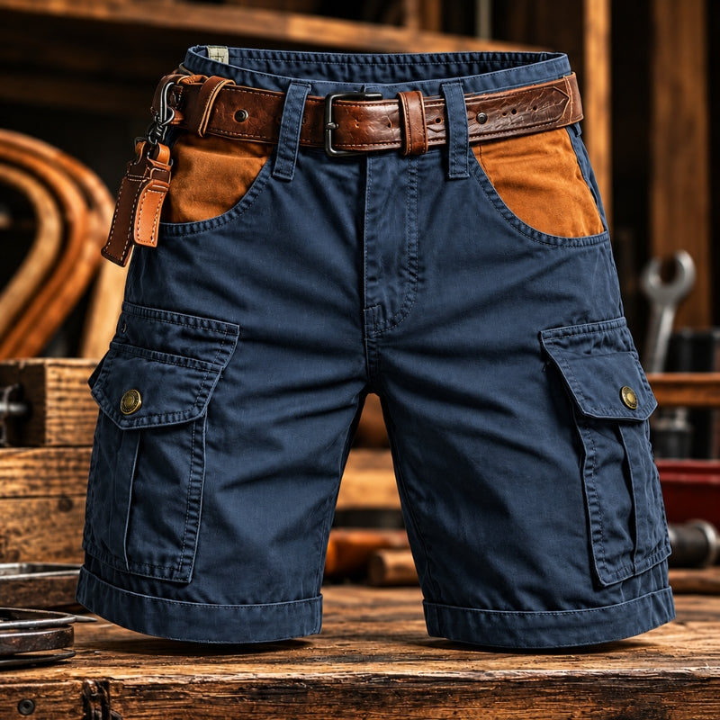 A pair of navy blue cargo shorts with brown leather accents, including a matching belt and a leather belt loop, displayed on a rustic wooden workbench with a workshop background.