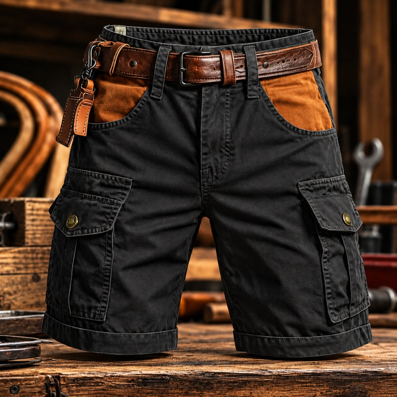 Black cargo shorts with brown leather accents, including a matching belt, belt loops, and a leather key fob, displayed on a rustic wooden workbench with a workshop background.