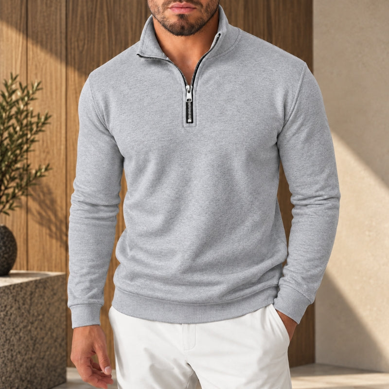 A man wearing a light gray heathered quarter-zip pullover sweater with white pants, standing in front of a warm wood-paneled wall.