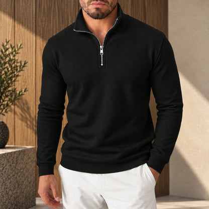 A man wearing a black long-sleeve quarter-zip pullover sweater with a stand-up collar and white pants, standing in front of a warm wood-paneled wall.
