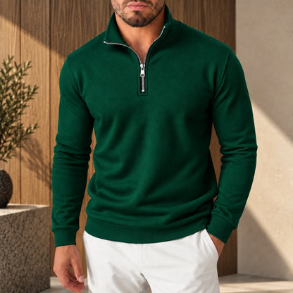 A man wearing a dark green long-sleeve quarter-zip pullover sweater with a silver zipper and white pants, standing in front of a wooden wall and concrete planter.