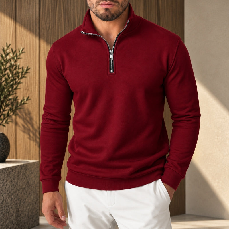 A man wearing a burgundy long-sleeve quarter-zip pullover sweater and white pants, standing in a modern indoor setting with wood paneling and soft sunlight.