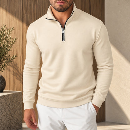 A man wearing a cream-colored long-sleeve quarter-zip pullover sweater with a black zipper pull, paired with white pants, standing in a modern indoor setting with warm sunlight and wood paneling.
