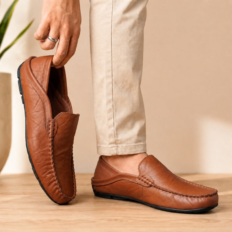 Brown leather loafers being held by a person wearing beige pants on a wooden floor.
