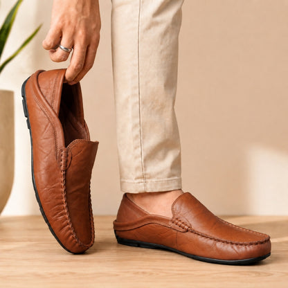 Brown leather loafers being held by a person wearing beige pants on a wooden floor.