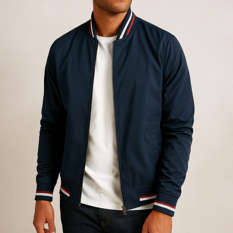 A man wearing a navy blue zip-up bomber jacket with red and white striped ribbed collar, cuffs, and hem, layered over a white t-shirt and dark jeans, against a plain light beige background.