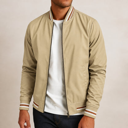 A man wearing a beige zip-up bomber jacket with brown and white striped ribbed trim on the collar, cuffs, and hem, layered over a white t-shirt and dark jeans.