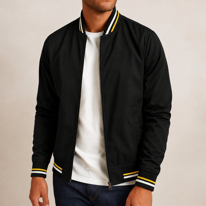 A man wearing a black zip-up bomber jacket with yellow and white striped ribbed collar, cuffs, and hem, layered over a white t-shirt and dark blue jeans, set against a plain beige background.