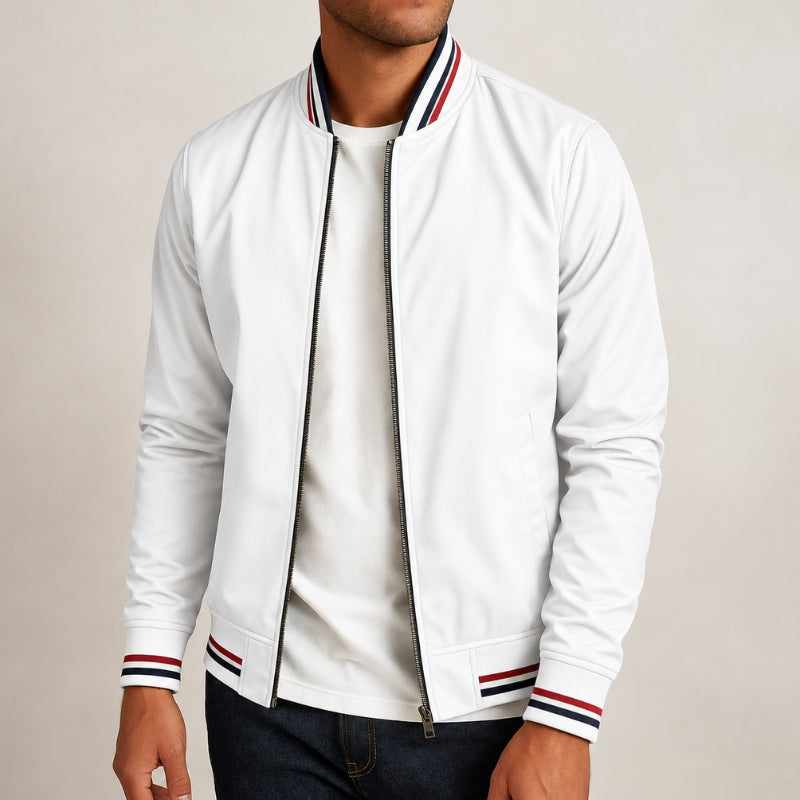 A man wearing a white zip-up bomber jacket with red and navy striped ribbed collar, cuffs, and hem, paired with a white t-shirt and dark jeans, against a plain light beige background.
