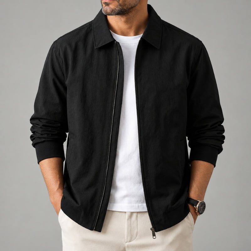 A man wearing a black zip-up jacket with a collar over a white t-shirt and light beige trousers, shown from the chin down against a solid gray background.