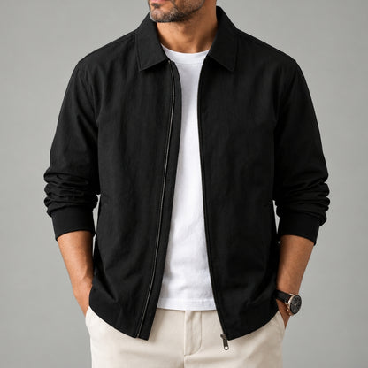 A man wearing a black zip-up jacket with a collar over a white t-shirt and light beige trousers, shown from the chin down against a solid gray background.