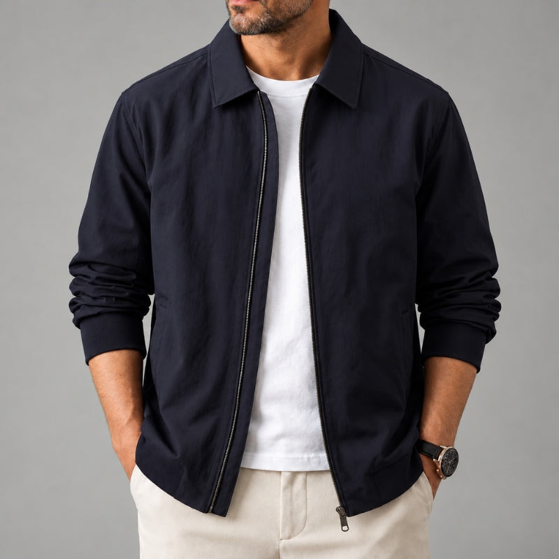 A man wearing a dark navy blue zip-up jacket over a white t-shirt and beige trousers, standing against a solid gray studio background.
