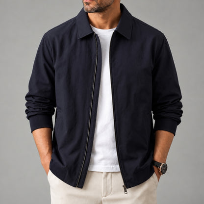 A man wearing a dark navy blue zip-up jacket over a white t-shirt and beige trousers, standing against a solid gray studio background.