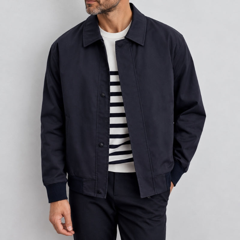 A man wearing a dark navy blue jacket over a white and navy striped t-shirt, paired with dark trousers, photographed against a light gray studio background.