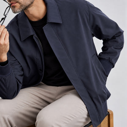 A man wearing a navy blue lightweight jacket over a black shirt and beige trousers, seated against a light gray background.