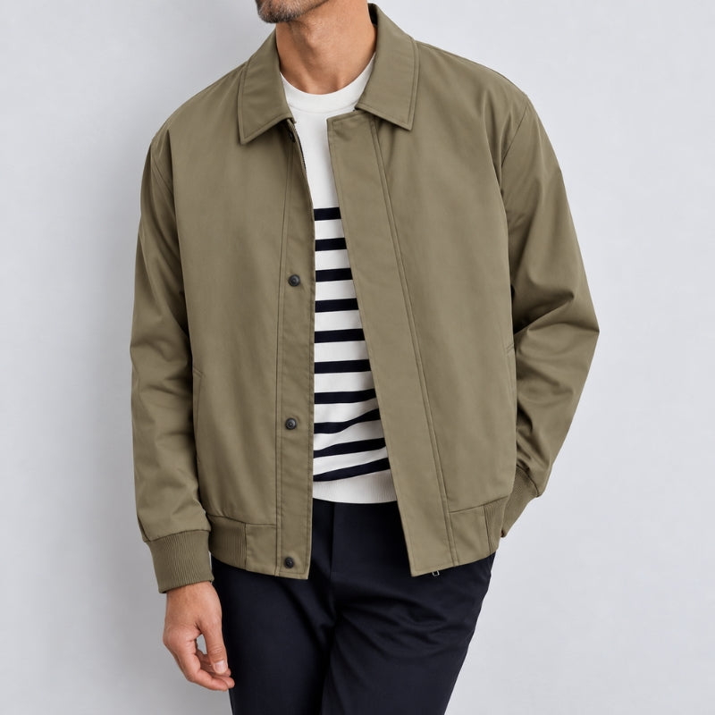 A man wearing an olive green utility jacket over a white and navy striped t-shirt and dark trousers, photographed against a light gray studio background.